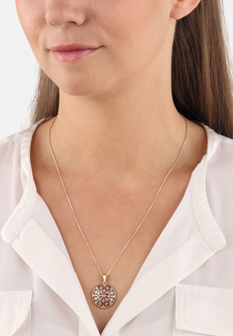 Naszyjnik z wisiorkiem w kolorze różowego złota, o okrągłym kształcie z misternymi wzorami wyciętymi w metalu, zawieszony na cienkim łańcuszku, noszony na białej bluzce.