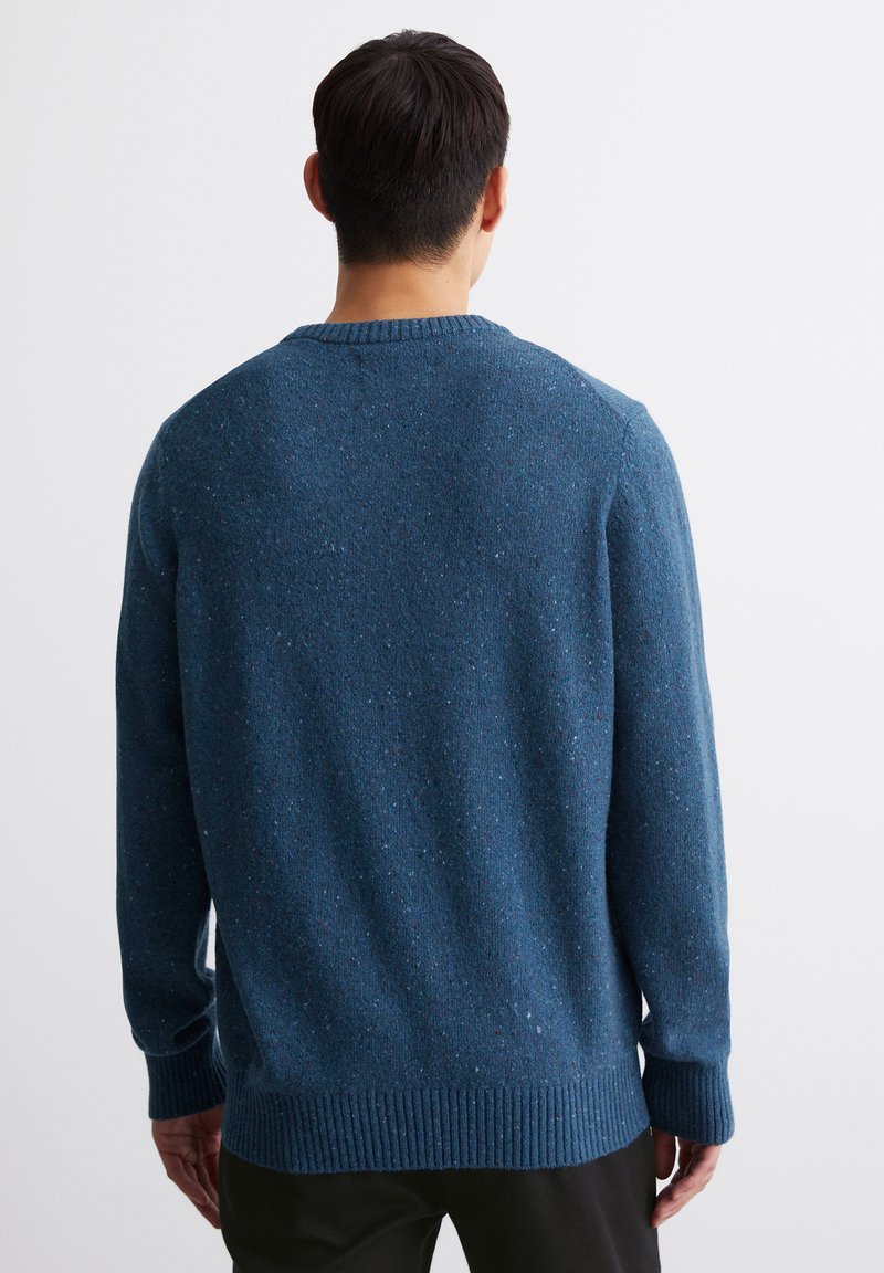 Mann, der von hinten zu sehen ist, trägt einen blauen strukturierten Strickpullover und schwarze Hosen vor einem schlichten weißen Hintergrund.