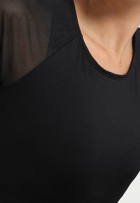 Lähikuva henkilöstä, joka pukeutuu mustaan paitaan, jossa on pyöreä kaula-aukko ja läpikuultavat verkkohihat harteilla.