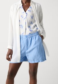 Blouse blanche portée sur une chemise à motif floral et un short bleu clair ; comporte un bouton et une taille élastique. Le tissu semble léger et doux.