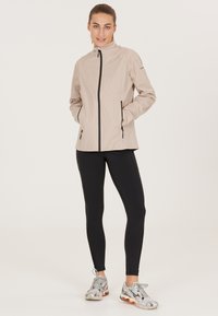 Lys beige vindjakke med frontlynlås, sidelommer og høj krave. Sorte detaljer langs syningerne. Båret med sorte tights og sneakers.