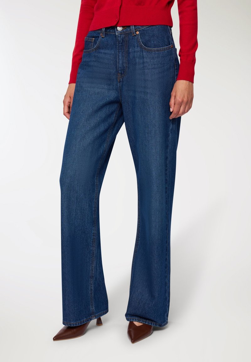 Jeans a gamba larga in denim blu scuro. Presentano cinque tasche, cuciture arancioni e una vita alta. Indossati con un maglione rosso e tacchi marroni.