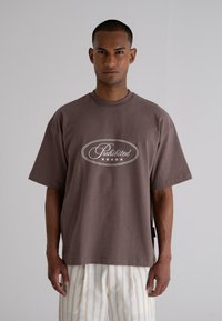 Brown oversized cotton t-shirt featuring a white oval 'Prohibited' graphic and star motif; model wears striped light-colored pants.