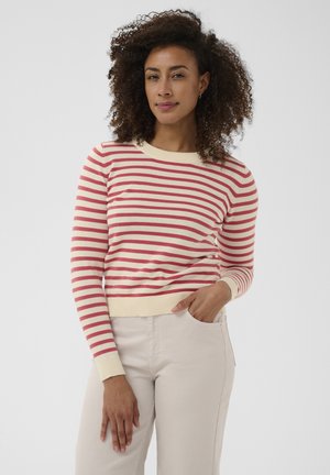 Femme aux cheveux bouclés portant un pull à manches longues rouge et blanc à rayures et un pantalon beige clair, debout avec une main dans la poche.