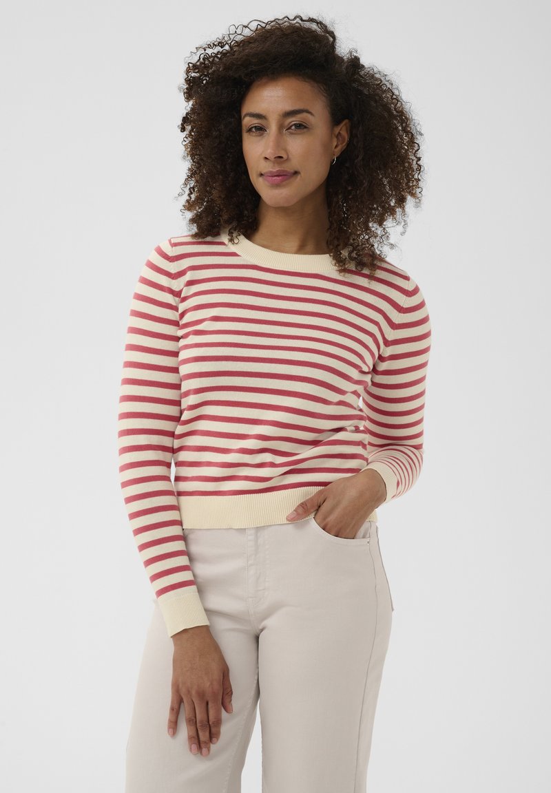 Femme aux cheveux bouclés portant un pull à manches longues rouge et blanc à rayures et un pantalon beige clair, debout avec une main dans la poche.