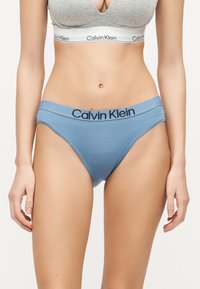 Femme portant un soutien-gorge de sport gris Calvin Klein et un sous-vêtement bleu Calvin Klein, debout, les bras détendus le long du corps, sur un fond uni.