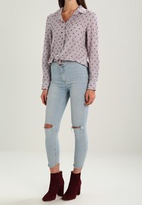Camicia button-up a maniche lunghe con motivo rosso e blu, abbinata a jeans skinny chiari strappati e stivali alla caviglia bordeaux.