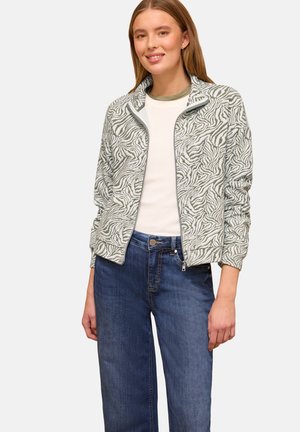 Ung kvinde iført en lysegrå og hvid zebra-print lynlåstrøje, hvid skjorte og blå jeans, smilende med hænderne afslappet ved siden.