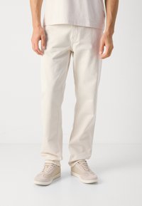 Man wearing off-white straight-leg pants and matching sneakers, standing on a white floor against a plain background.