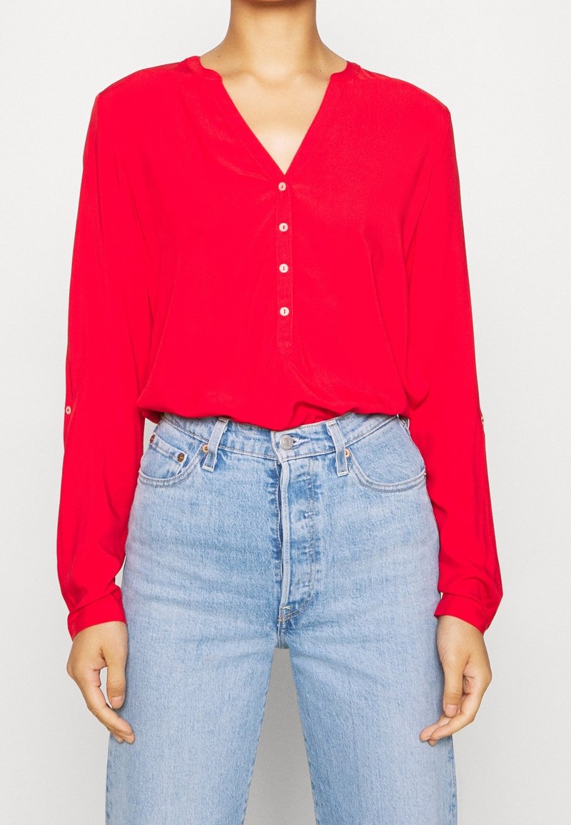 Frau trägt eine leuchtend rote Bluse mit langen Ärmeln und Knöpfen sowie hellblaue hochtaillierte Jeans und steht vor einem einfarbigen Hintergrund.
