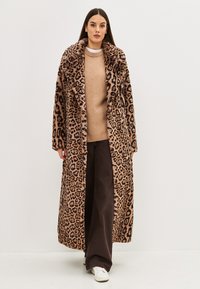 Abrigo de piel sintética de leopardo con cuello ancho, mangas largas y apertura frontal, llevado sobre un suéter beige y pantalones de pierna ancha marrones.