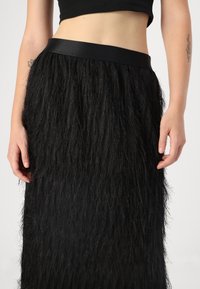 Black skirt with textured fringe design, featuring a smooth waistband. The material appears soft and flowing, adding depth to the silhouette.