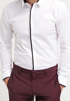 Homme portant une chemise blanche ajustée avec un liseré noir et un pantalon bordeaux, mains dans les poches, debout devant un fond neutre.