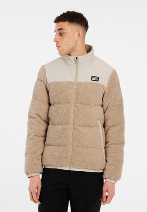Chaqueta de forro polar beige con un panel superior ligero en beige, que cuenta con cierre de cremallera, bolsillos laterales y un parche con el logo en el pecho.