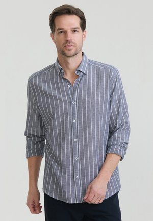 Férfi, aki szürke hosszú ujjú gombos ingben, fehér függőleges csíkokkal, feltűrt ujjakkal, áll egy egyszínű szürke háttér előtt.