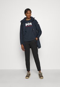Chaqueta impermeable azul marino con capucha, combinada con una camiseta oscura con un logo blanco y rojo, pantalones negros y zapatillas marrones.