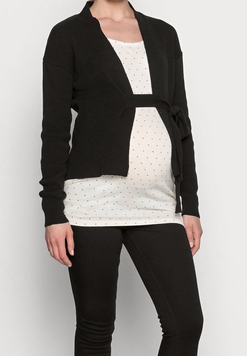 Czarny prążkowany kardigan z długimi rękawami, z paskiem związanym w talii, noszony na górze w białe kropki i czarne spodnie.