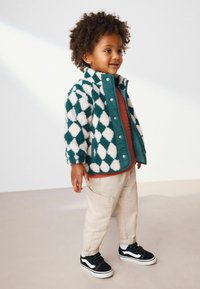 Veste en polaire à motif losange vert et blanc, pantalon cargo beige, baskets noires avec accents blancs, tissu texturé, coupe décontractée.