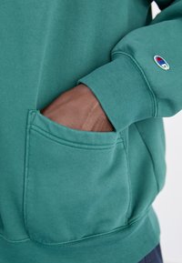 Ciemnoskóra dłoń w kieszeni przedniej bluzy Champion w kolorze turkusowym, z widocznym logo na rękawie, na jasnym tle.