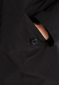 Abrigo negro con cierre de botones, bolsillo lateral y una textura suave y mate. La tela parece ser duradera y resistente al agua.