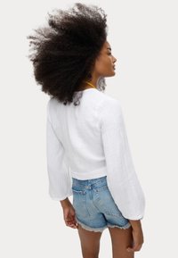 Top bianco leggero a maniche lunghe con tessuto texture. Abbinato a pantaloni corti in denim chiaro con orli sfrangiati. Modello visto di spalle.
