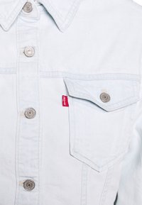 Hellblaues Denim-Jackett mit silbernen Druckknöpfen, spitzem Kragen und einer Vordertasche mit einem kleinen roten Levi's-Label. Strukturiertes Material.