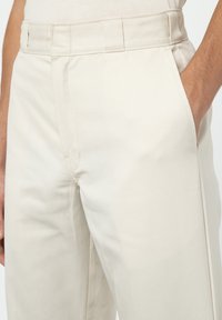 Béžové kalhoty s hladkým bavlněným materiálem, mají rovný střih, boční kapsy a plochý přední design. Jsou viditelné detaily šití.