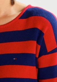 Czerwono-granatowy sweter w paski o prążkowanej strukturze i okrągłym dekolcie. Posiada małe logo haftowane blisko dolnego obszycia.