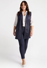 Marineblaue ärmellose Weste aus Kunstleder mit Seitentaschen, kombiniert mit einem weißen Hemd mit Knöpfen und passenden marineblauen Hosen sowie hellen Schuhen.