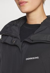 Svart værbestandig jakke med glidelås foran, trykknapper og hette. Har hvit "Didriksons" logo på venstre brystområde.