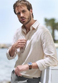 Beige gestreiftes Langarmhemd mit Knopfkragen, kombiniert mit einer blauen Uhr an einem braunen Lederarmband und hellgrauen Hosen.