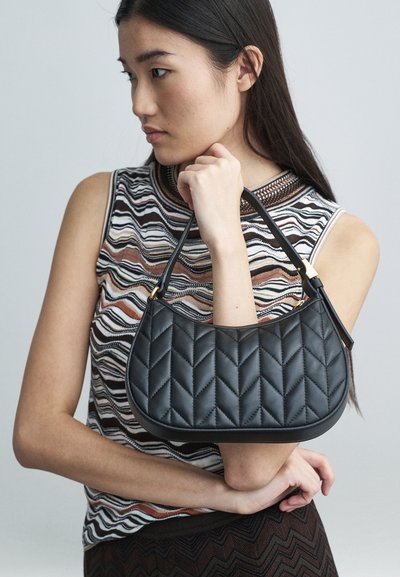 Νέα γυναίκα με αμάνικο μοτίβο που κρατά μια μαύρη διακοσμημένη τσάντα με κοντό λουρί σε ένα απλό φόντο.