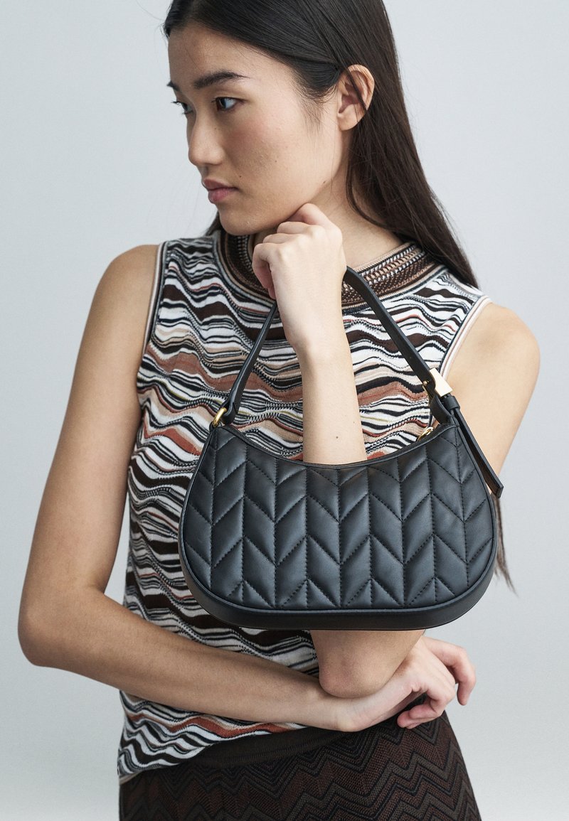 Giovane donna in top senza maniche con motivo, che tiene una borsa a tracolla nera trapuntata, davanti a uno sfondo neutro.