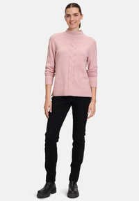 Helles rosa Strickpullover mit hohem Kragen, versehen mit einem zentralen Zopfstrickmuster. Kombiniert mit schwarzen Slim-Fit-Hosen und schwarzen Stiefeletten.