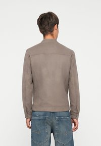 Chaqueta de ante gris con cuello, mangas largas y detalles de botones en los puños. Presenta un corte entallado y se lleva con pantalones de mezclilla azul.