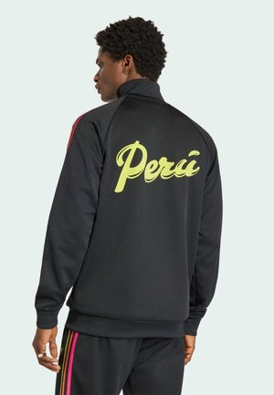 Mand iført sort træningsjakke og bukser med pink og gule striber, "Percé" trykt i gult på jakke ryg, stående i profil.