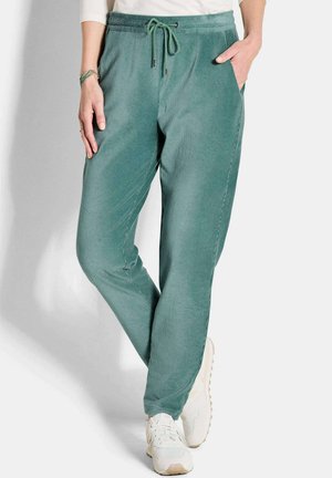 Person trägt tealgrüne Cord-Jogginghosen mit Kordelzug in der Taille und weiße Sneakers, steht mit einer Hand in der Tasche.