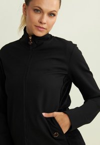Giacca nera con zip e colletto alto, realizzata in un tessuto liscio, dotata di tasche laterali e un dettaglio dorato vicino all'orlo. Pannelli laterali strutturati.