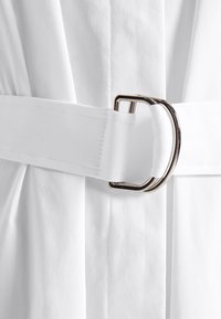 Cintura di tessuto bianco con una grande fibbia di metallo argentato, caratterizzata da una texture liscia e da un design ampio e piatto. La cintura è stretta in vita.