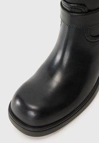 Botín de cuero negro hasta el tobillo con punta redondeada, textura suave y correa ajustable. Suela resistente con un discreto detalle de costura.