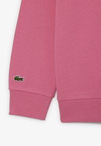 Felpa rosa in tessuto con polsini a costine; presenta un piccolo logo di coccodrillo ricamato in verde sul polsino sinistro. Texture liscia, design casual.