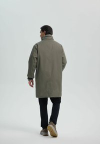 Oliwkowy, wodoodporny płaszcz z wysokim kołnierzem, sięgający do połowy ud. Posiada luźny krój oraz długie rękawy, noszony z ciemnymi spodniami i butami.