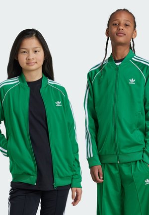 Des vestes zippées vertes avec des rayures blanches sur les manches, fabriquées en tissu doux. Ils portent tous les deux des chemises noires ; l'un porte un pantalon vert assorti.