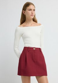 Jonge vrouw met lang blond haar, gekleed in een witte off-shoulder ribbeltop en een hoog opgesneden rode textuurrok met een gouden knoop.