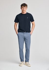 Svart bomullströja med ett litet emblem på bröstet, matchad med ljusblå chinos och krämfärgade sneakers, stående mot en neutral bakgrund.