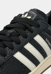 Μαύρο αθλητικό παπούτσι με υφή και μοτίβο που μοιάζει με δέρμα φιδιού, με λευκές ρίγες, μαύρα κορδόνια και ετικέτα με λογότυπο στη γλώσσα.