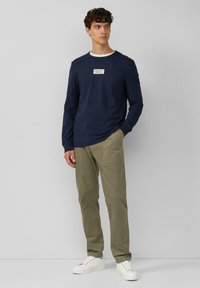 Maglietta a maniche lunghe blu navy con etichetta bianca sul petto, abbinata a pantaloni in velluto a coste verde oliva e sneakers bianche. Design semplice e casual.
