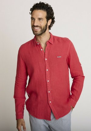 Homme souriant aux cheveux bouclés foncés et à la barbe, portant une chemise rouge à manches longues boutonnée et un pantalon gris clair, debout devant un fond uni.