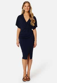 Námořnická modrá zavinovací šaty z jemného, elastického materiálu. Mají výstřih do V, krátké rukávy typu batwing a délku ke kolenům s bočním rozparkem.