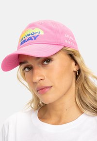Gorra de algodón rosa con visera curva, que presenta un gráfico de "BYRON BAY" en azul y amarillo. Branding adicional en el lateral en blanco.